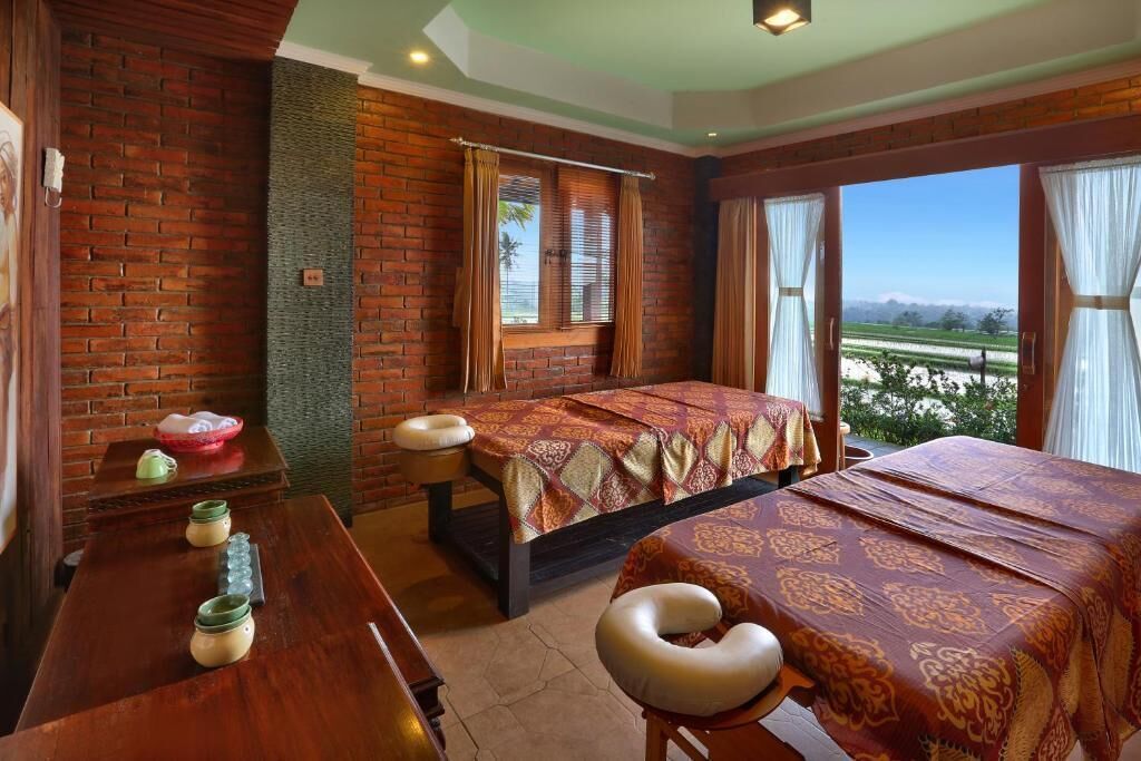 Habitación de hotel con pared de ladrillo, cama con colcha estampada, escritorio con un tazón y frascos, y vista al paisaje a través de la ventana.