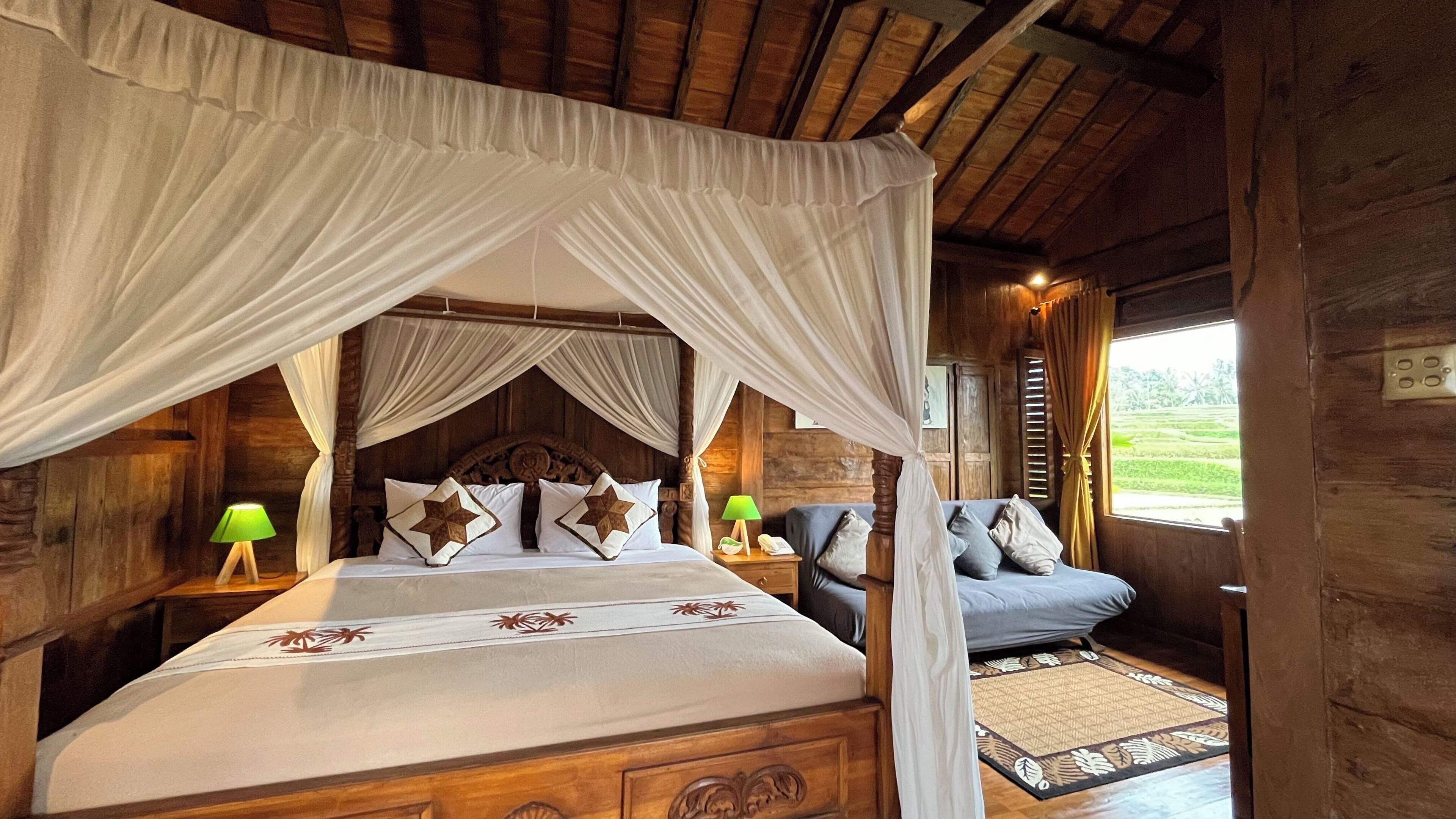 Una cama con dosel de madera, una mesita de noche con lámpara, y vista a un paisaje verde a través de una ventana.