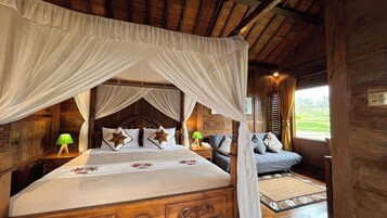 A wooden four-poster bed with a canopy, a bedside table with a lamp, and a view of a green landscape through a window.