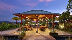 An outdoor dining area with a wooden pergola, tables, and benches, surrounded by lush greenery and a view of a body of water. - Ti Amo Bali (Penebel)