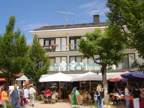 An outdoor cafe area with tables and umbrellas, a building with multiple signs including 'Cafe Bistro', and a flag display. - Gästehaus Café Heck (Titisee-Neustadt)