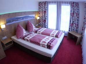 Ein Doppelbett mit karierter Decke, zwei Kissen, ein Nachttisch, eine Wandlampe und ein Landschaftsblick durch ein Fenster mit floralen Vorhängen.