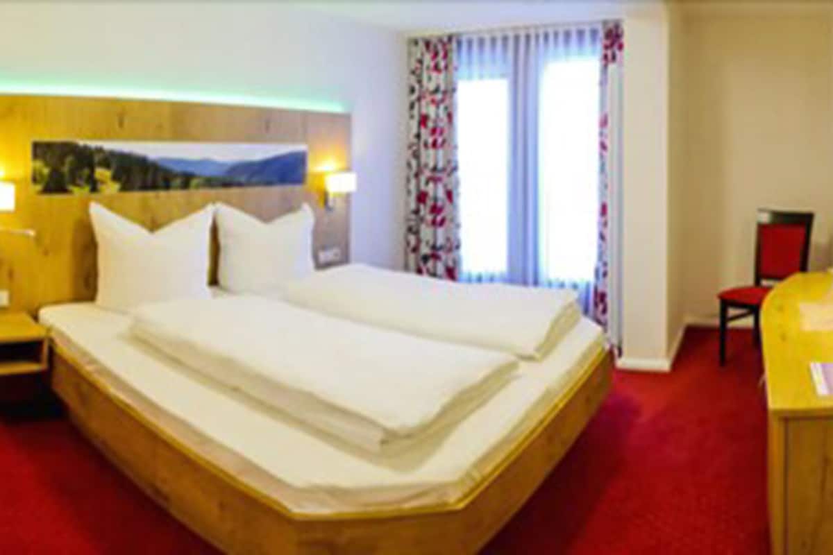 A hotel room with a double bed, wooden headboard, a small bedside table, a desk with a chair, and a TV on the wall.