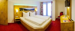 Ein Hotelzimmer mit Doppelbett, Holz-Kopfteil, einem kleinen Nachttisch, einem Schreibtisch mit Stuhl und einem an der Wand befestigten Fernseher.