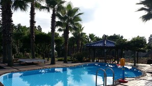 A swimming pool with a wooden deck, surrounded by palm trees and a gazebo. - Palace in the Forest Pension (Seogwipo)