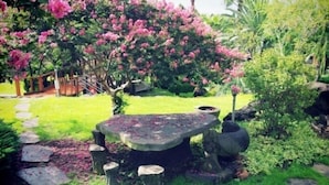 A garden with a stone table surrounded by chairs, a wooden deck, and flowering bushes. - Palace in the Forest Pension (Seogwipo)