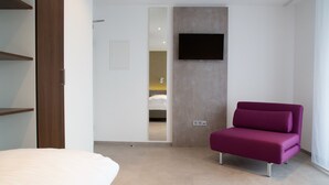 A modern hotel room with a bed, a purple sofa, a flat-screen TV mounted on the wall, and a door leading to a bathroom. - Apartmenthaus Renz (Aalen)