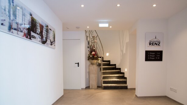 A modern hotel lobby with a staircase, a framed artwork on the wall, and a sign indicating room stages. - Apartmenthaus Renz (Aalen)