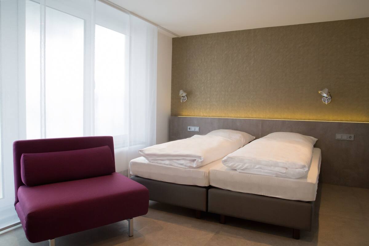 A modern hotel room with a bed, a red chair, and a large window.
