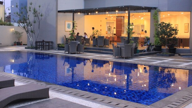 Une piscine avec des chaises longues, un espace de détente couvert et un bâtiment doté d’une façade en verre.