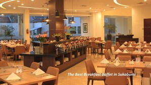 Un espace buffet pour le petit-déjeuner avec de nombreuses tables et chaises, un îlot central où les plats sont présentés, et de grandes fenêtres offrant une vue sur l’extérieur.