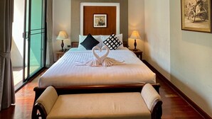 A hotel room with a large bed, a bench, a wooden ceiling, a framed picture on the wall, and a view through the glass door. - Yotaka Bangkok Hotel (Bangkok)