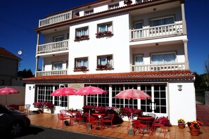 A two-story building with balconies, outdoor seating with red chairs and umbrellas, and flower arrangements. - Hotel Pipo (Sanxenxo)