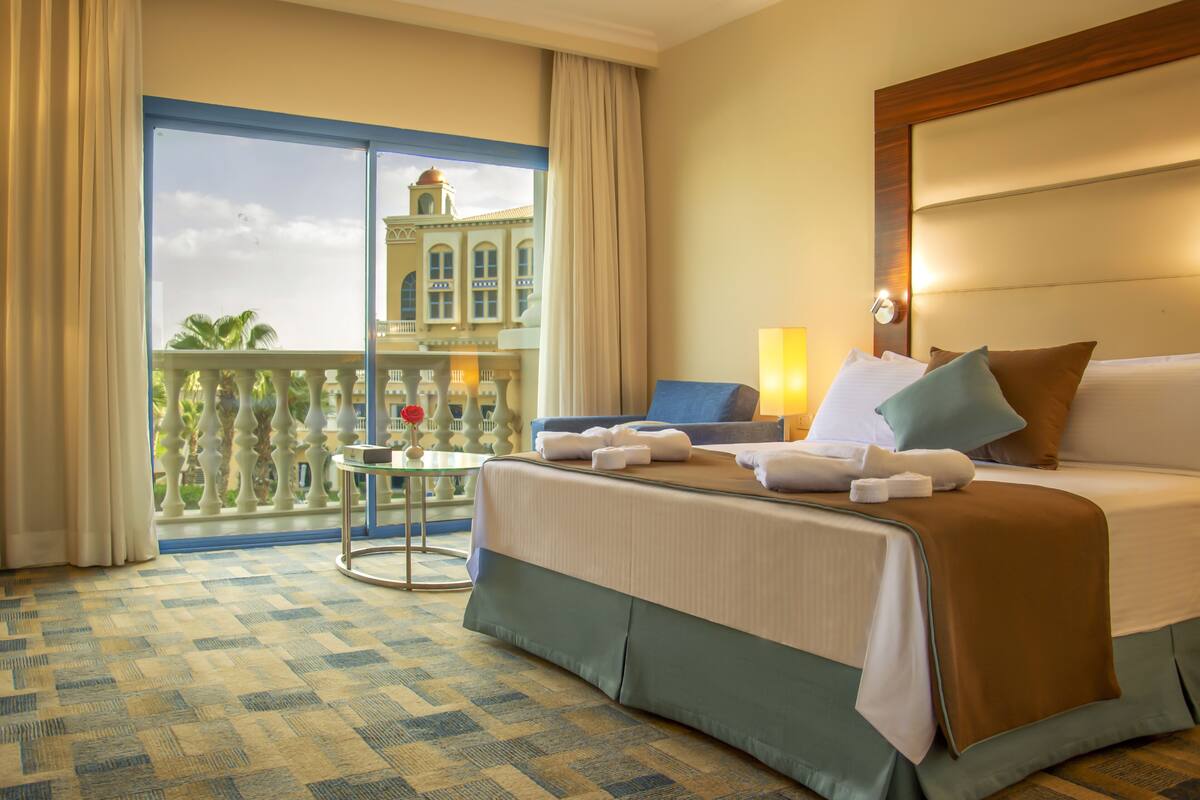 A hotel room with a large bed, a small table, a chair, and a balcony with a view of a building.