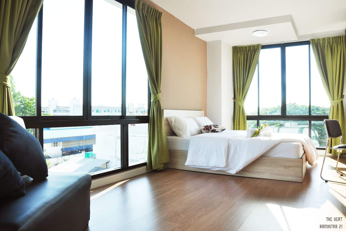 A modern hotel room with a large bed, two windows with green curtains, a view of the city, and a wooden floor.