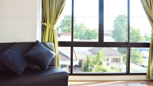 A room with a large window, a sofa with two cushions, and a view of buildings and trees outside. - The Vert Apartment (Bangkok)