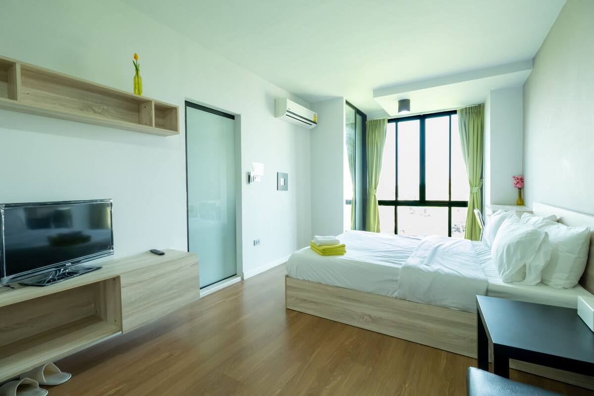 A modern hotel room with a bed, a TV, a desk, and a window with curtains.