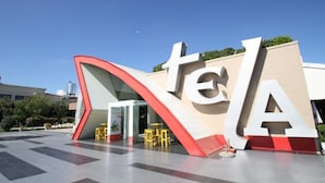A building with large letters spelling 'TELA' on the facade, a clear sky, and a paved area with yellow chairs. - Te Stela Resort (Kashar)