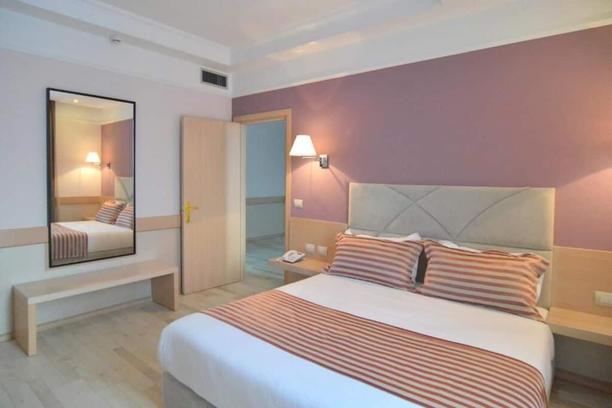 A hotel room with a bed, a bedside table, a lamp, a mirror, and a bench.