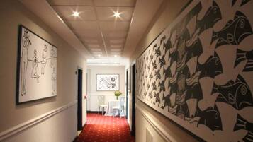 A hallway with a red carpet, framed artwork on the walls, and a dining area in the background.