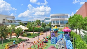 A playground with slides and climbing structures, surrounded by greenery and a building with a modern design. - Te Stela Resort (Kashar)