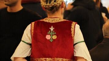 Une personne vêtue d’un costume traditionnel, avec un gilet rouge et une jupe verte, se tient dans un lieu intérieur bondé.