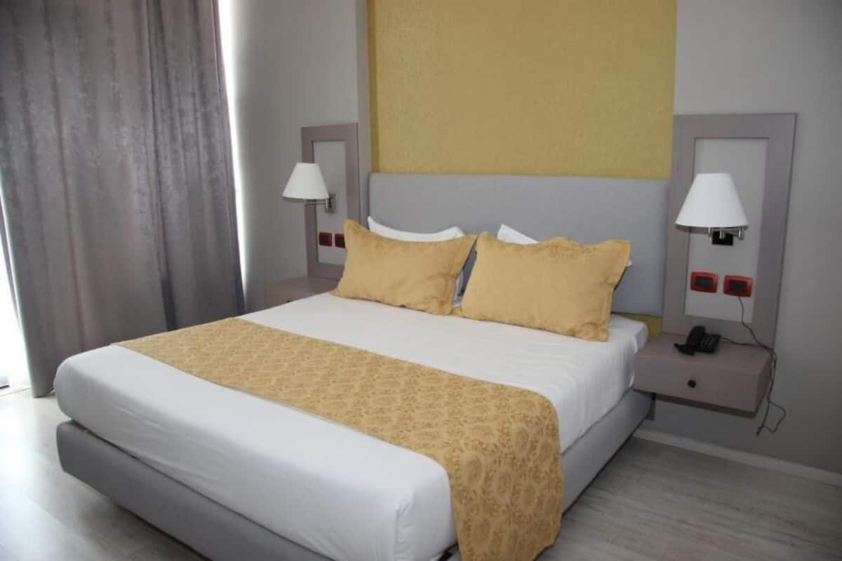 A hotel room with a bed, two bedside tables with lamps, a telephone, and a window with curtains.