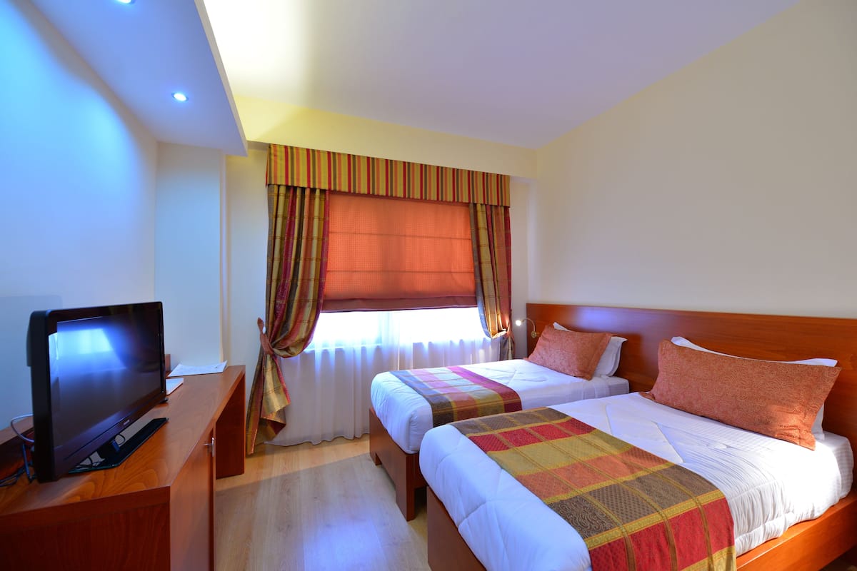 A hotel room with two beds, a TV on a wooden desk, and a window with curtains.