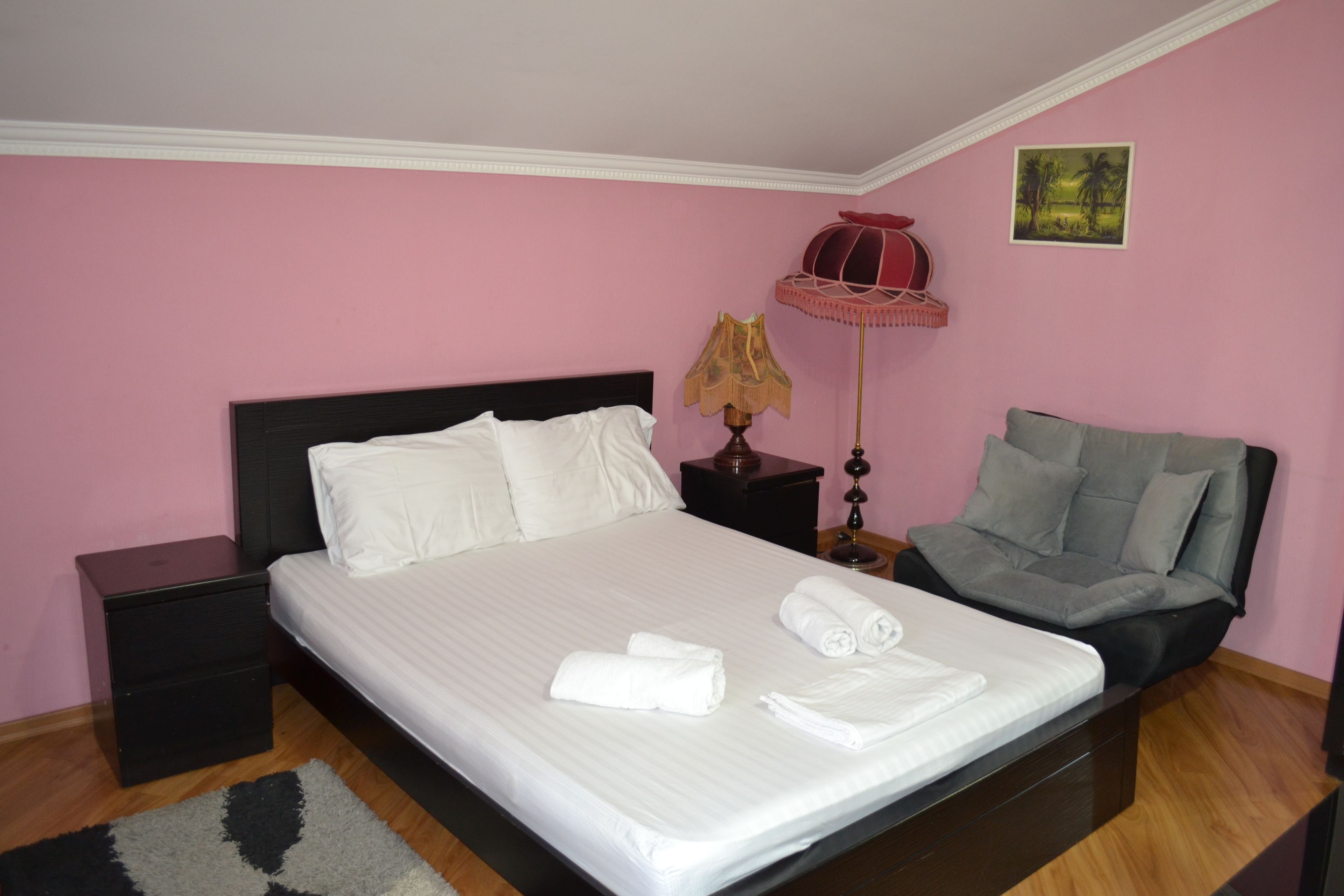 A neatly made bed with white linens and rolled towels, a gray sofa, a nightstand with a lamp, and a small table with a red lamp.