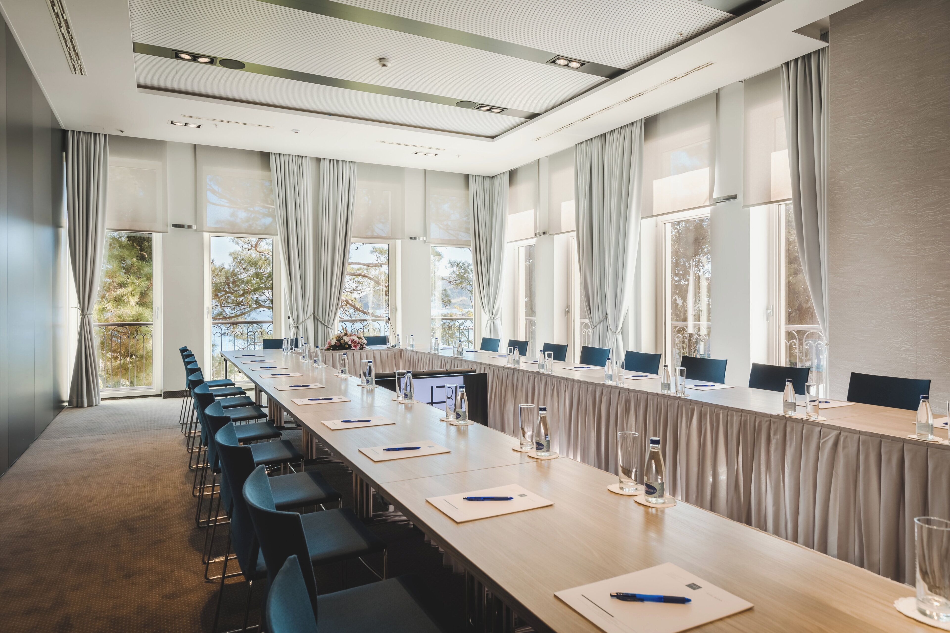 Une salle de conférence avec une longue table, des chaises et de grandes fenêtres équipées de rideaux.