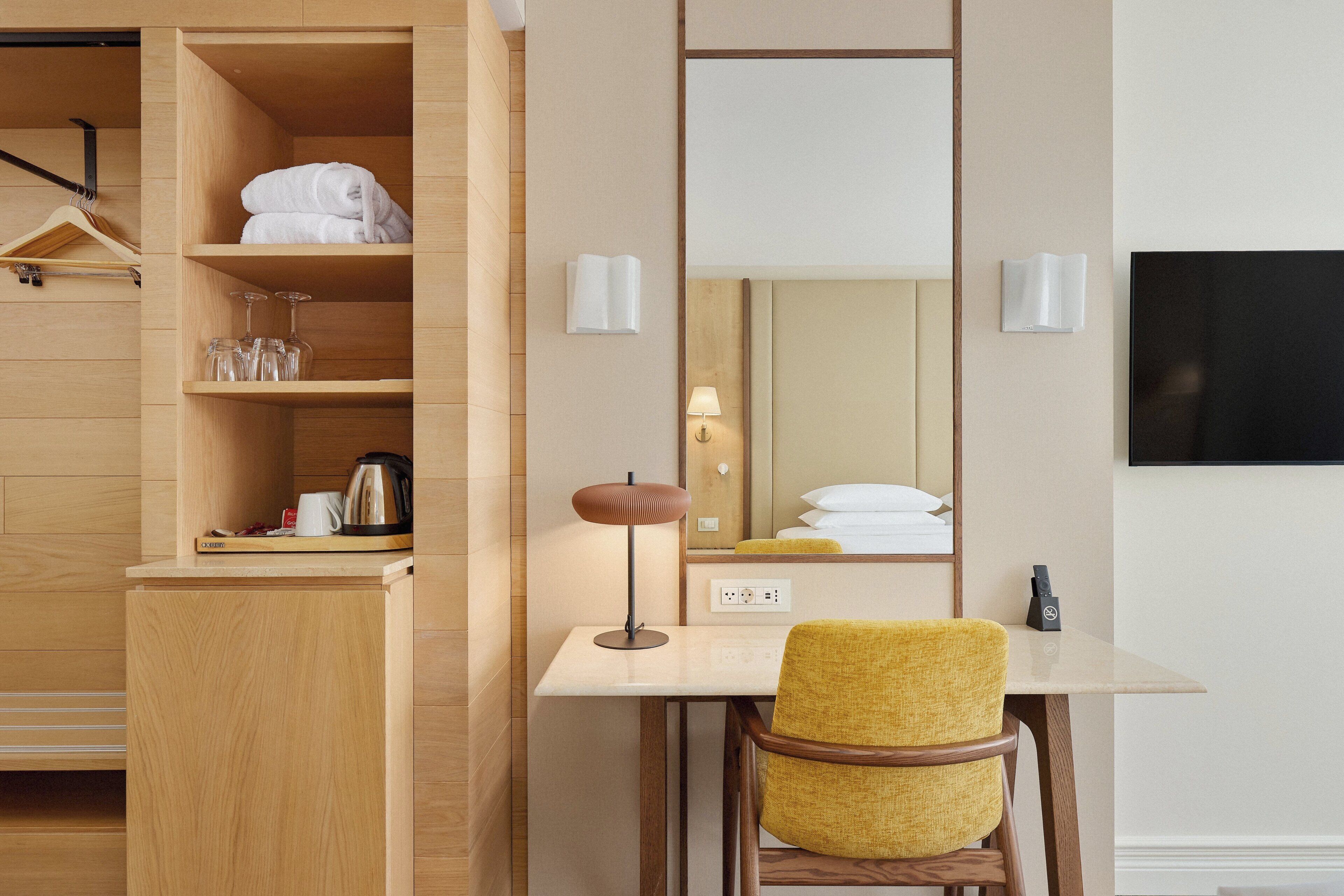 A hotel room with a wooden wardrobe, a desk with a lamp, a yellow chair, and a flat-screen TV.