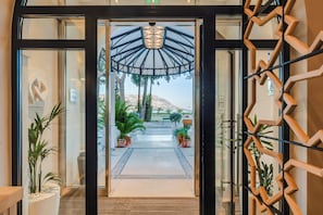 Entrada de hotel con puerta de cristal, separador geométrico de madera y plantas en macetas.