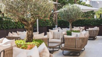 Un espace extérieur aménagé avec des chaises et des coussins en osier, un arbre et un parasol blanc.