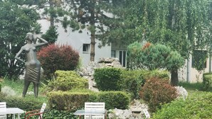 A serene outdoor seating area with tables and chairs, surrounded by lush greenery and a statue. - Hotel Slodes (Belgrade)