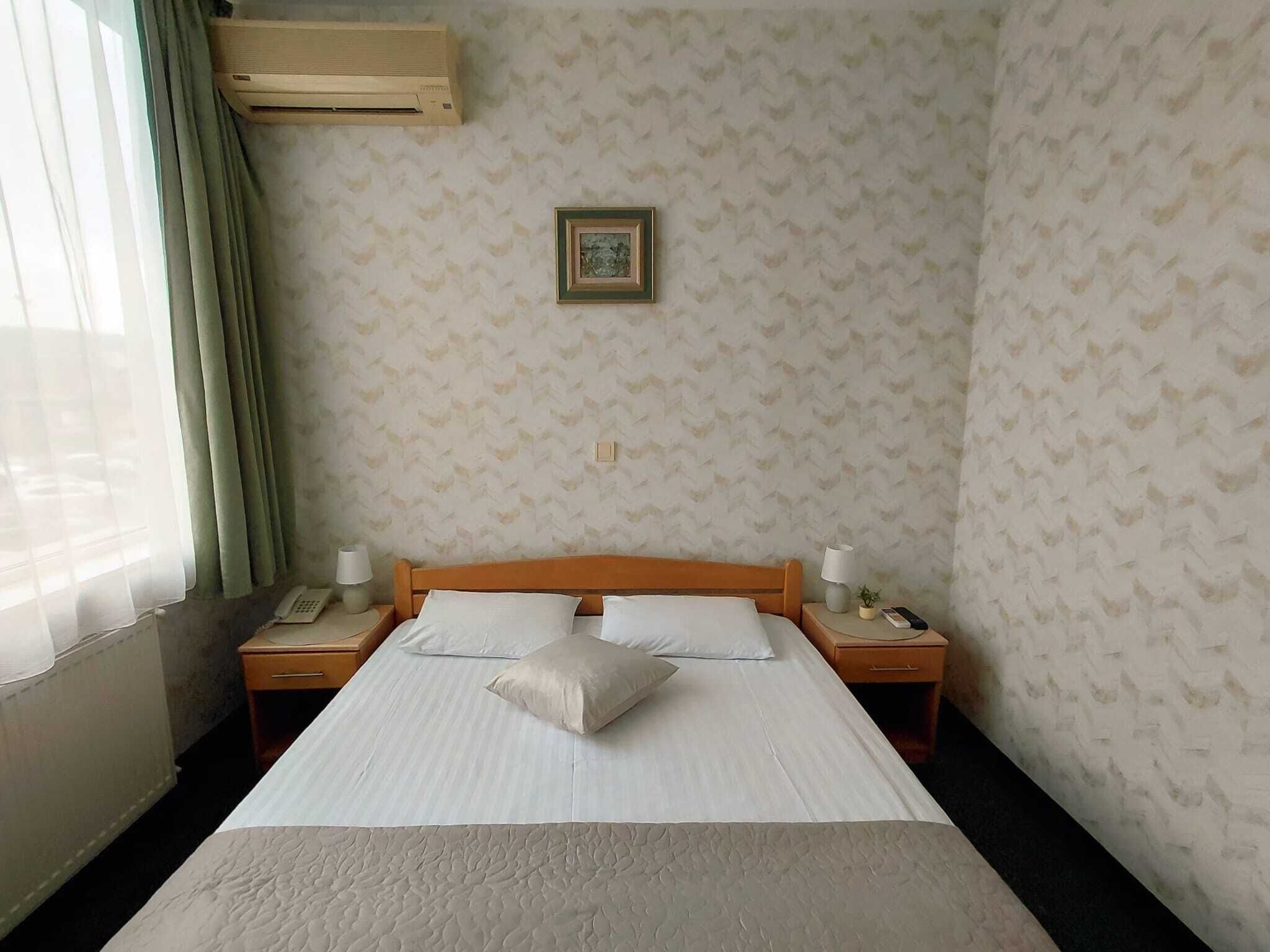 A hotel room with a bed, two bedside tables, a nightstand, a wall-mounted air conditioner, a window with curtains, and a patterned wallpaper.
