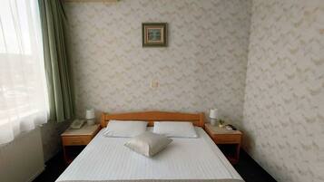 A hotel room with a bed, two bedside tables, a nightstand, a wall-mounted air conditioner, a window with curtains, and a patterned wallpaper.