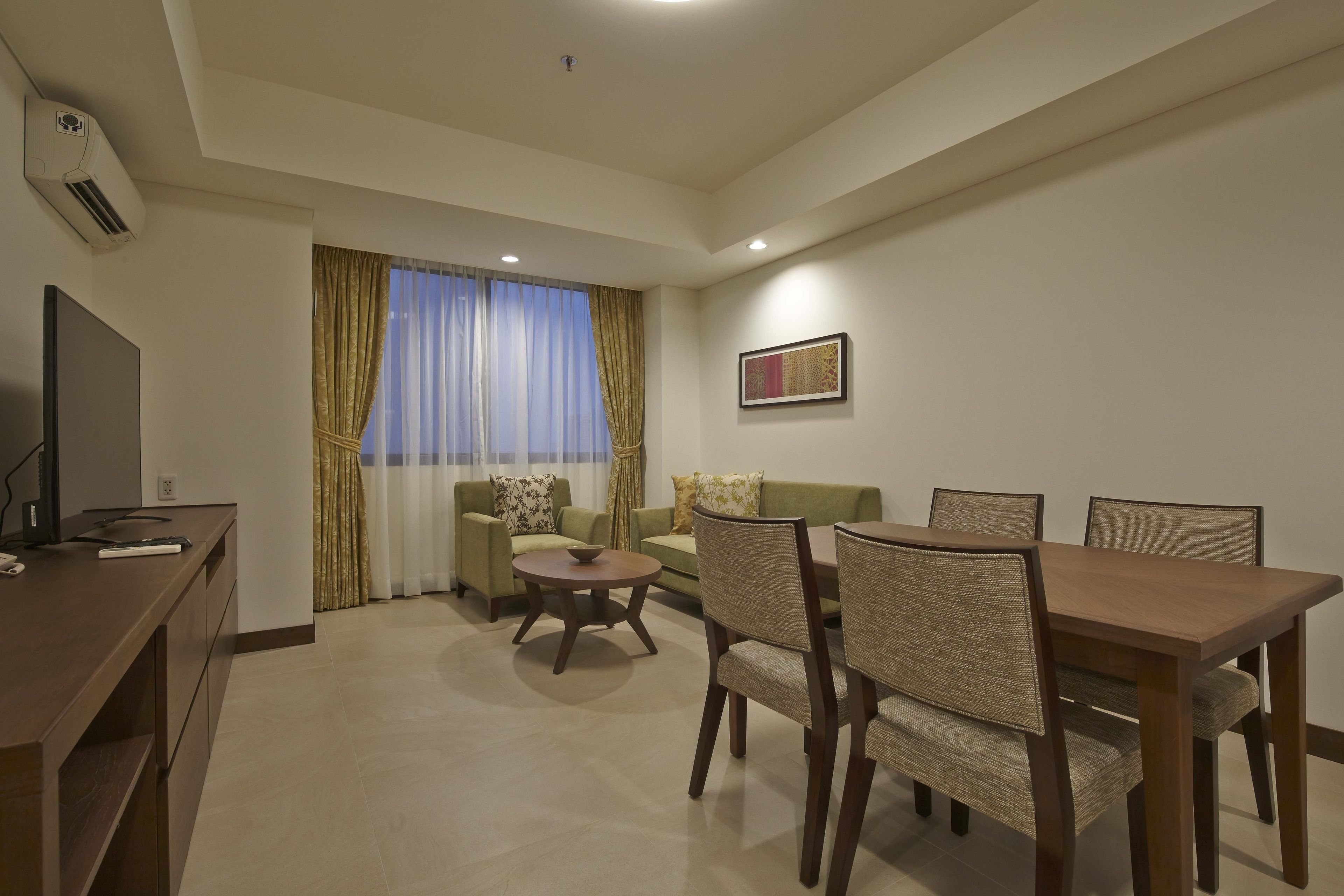 Una sala de estar con sofá, una mesa de comedor y sillas. Hay un televisor sobre un mueble bajo, una ventana con cortinas y un equipo de aire acondicionado en la pared.