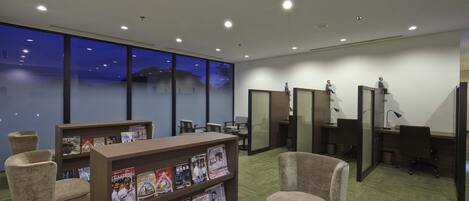A modern office space with a large bookshelf, comfortable seating, individual workstations, and large windows offering a view of the outside.