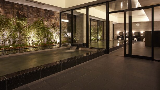 A modern indoor pool area with glass walls, a waterfall feature, and a brick wall with greenery.