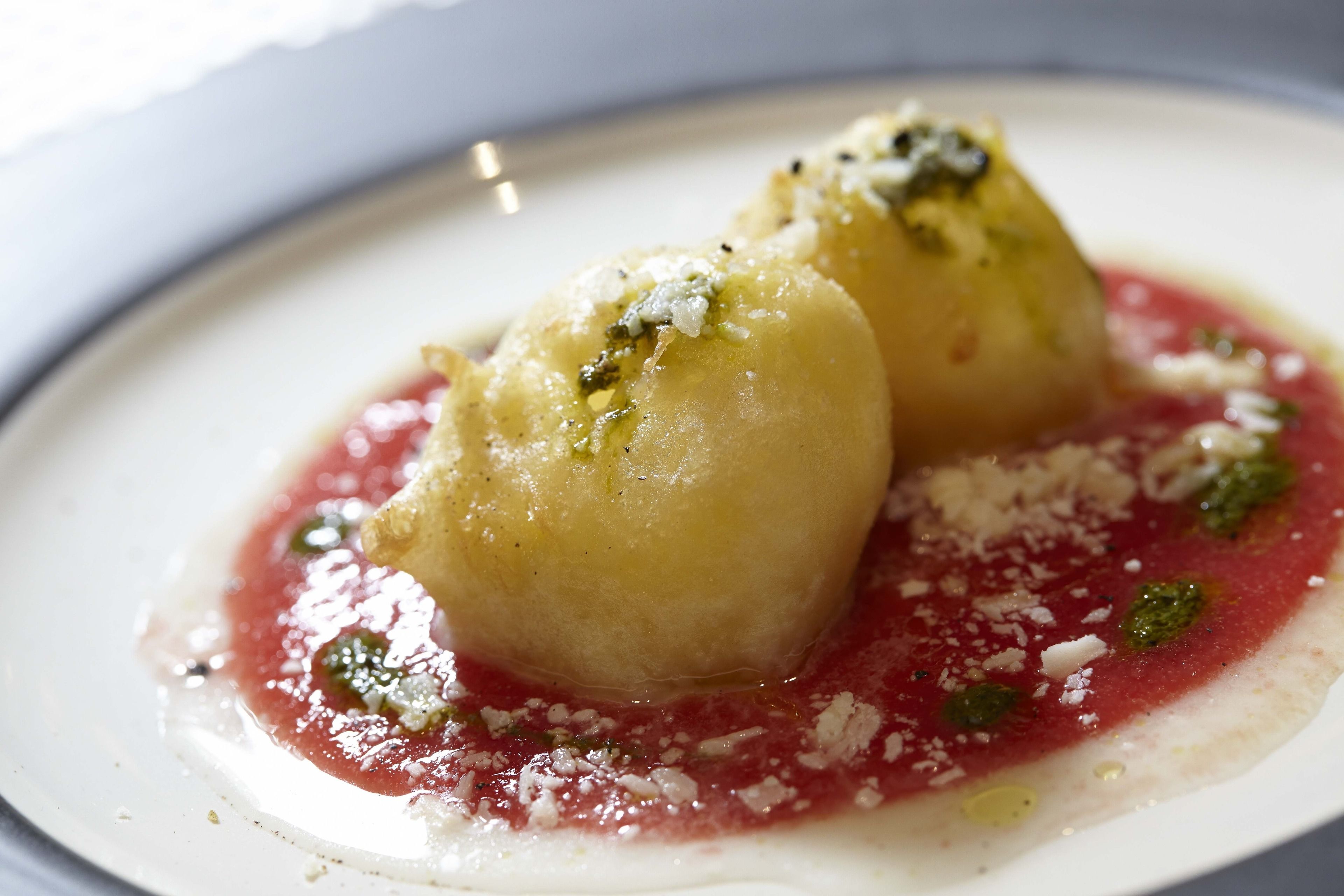 Un postre con dos bolitas redondas de color amarillo pálido sobre un plato blanco, cubiertas con queso rallado y salsa roja.