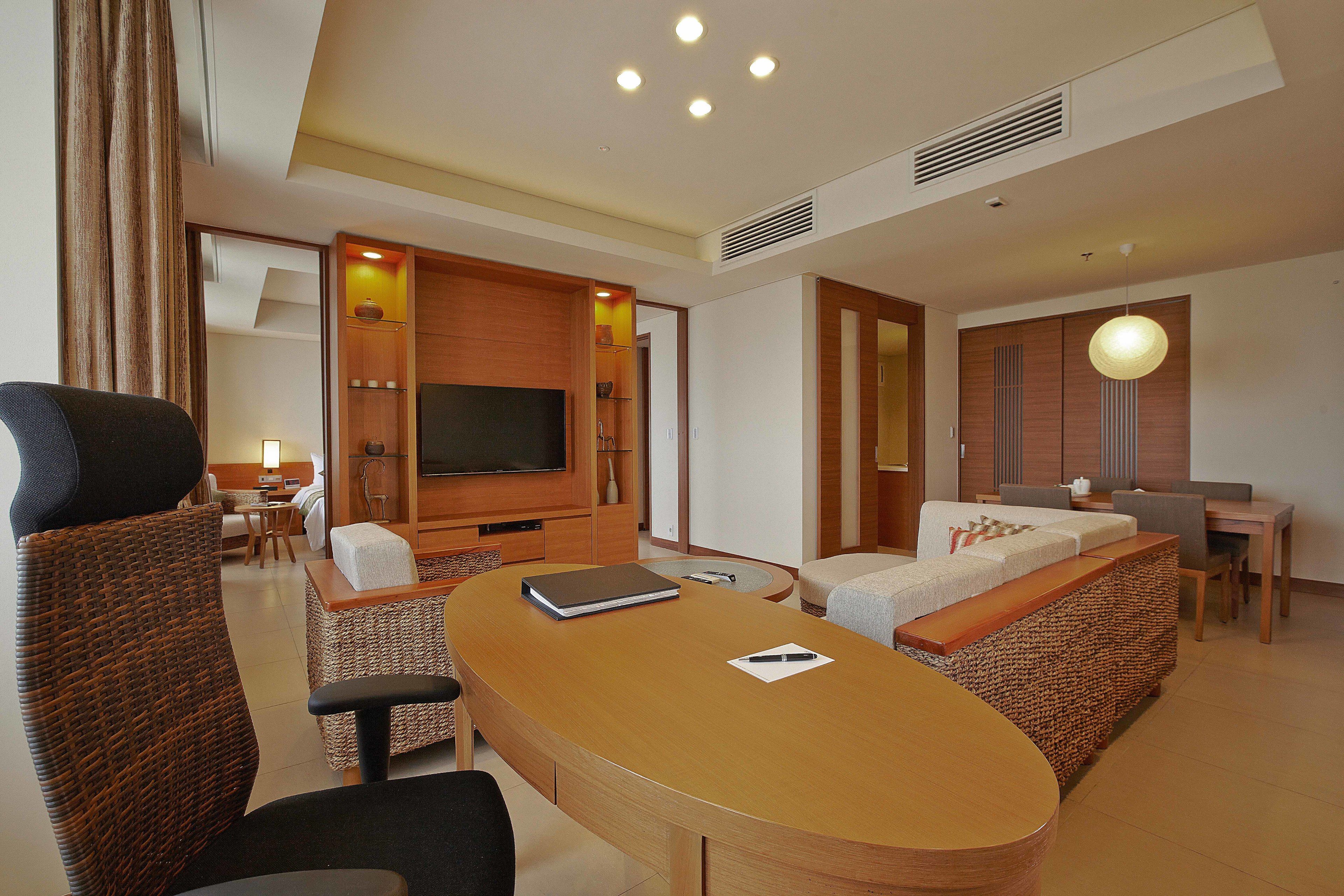 A modern hotel room with a wooden dining table, a sofa, a TV, and a separate dining area.