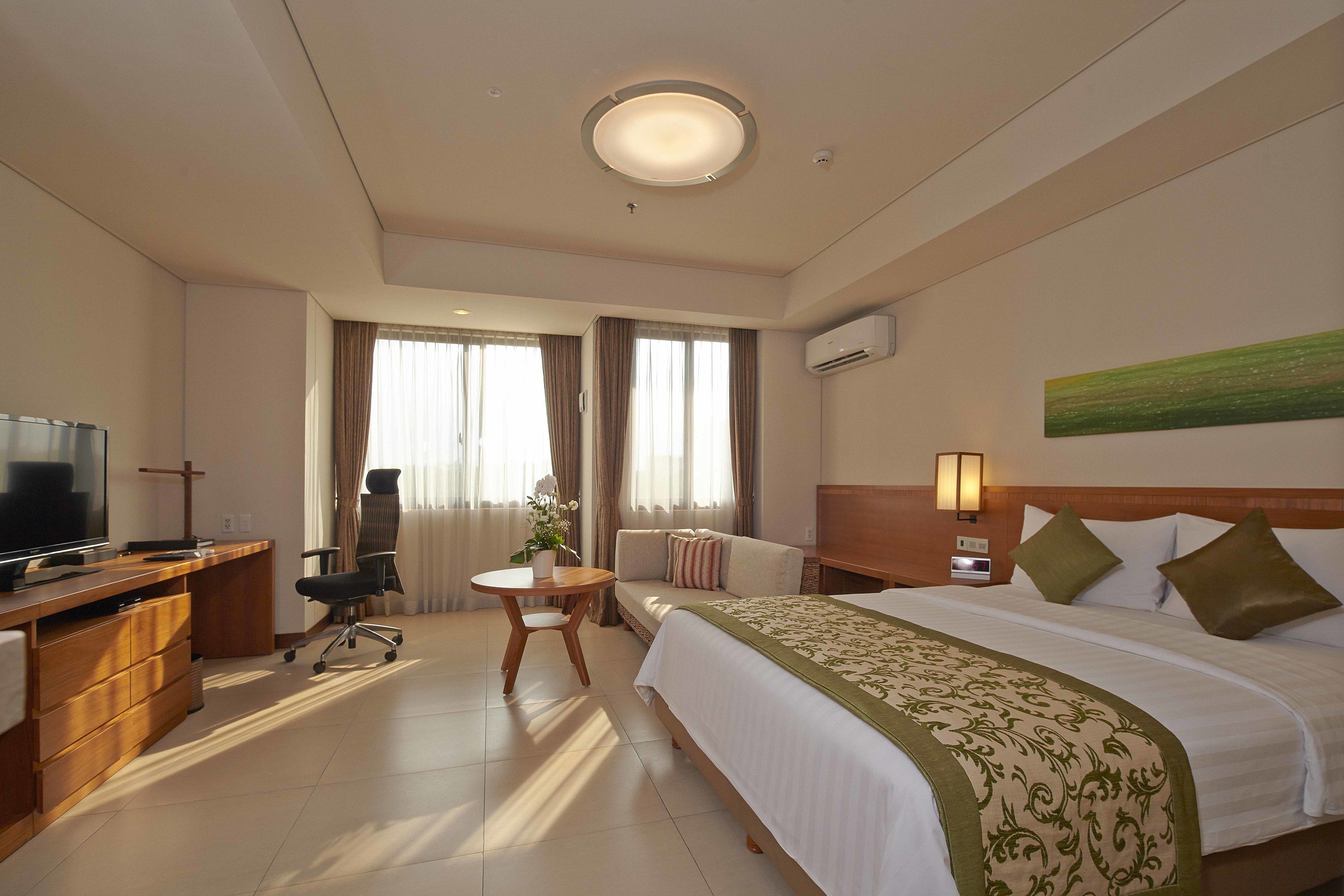 A hotel room with a bed, a desk, a chair, a TV, a small table, and a window with curtains.