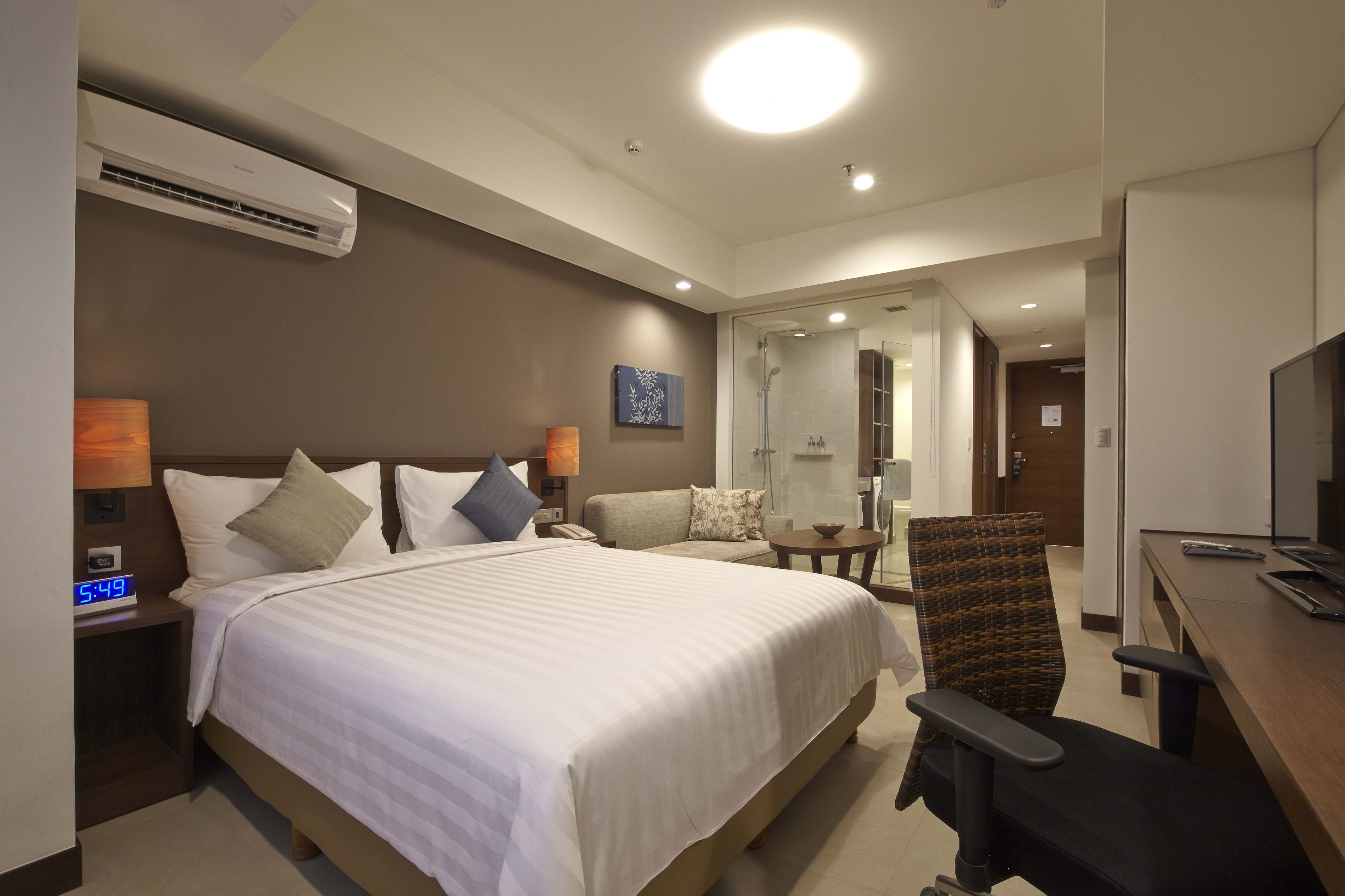 A hotel room with a large bed, a desk with a chair, a TV, and an air conditioning unit.