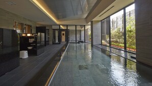 A modern indoor pool area with a glass barrier, a water feature, and a view of greenery outside.