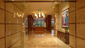 A hotel lobby with a glass entrance, a reception desk, and a large window offering a view of greenery.