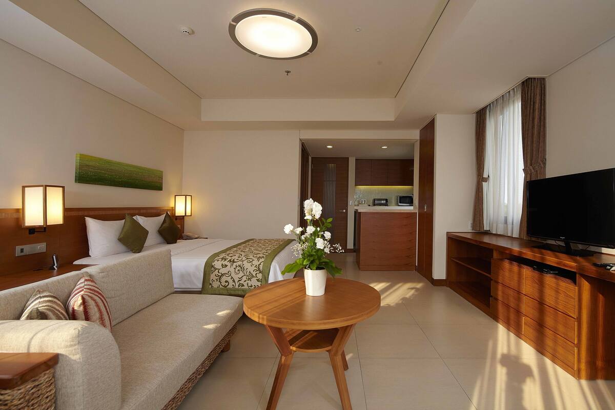 Een hotelkamer met een bank, een klein houten tafeltje, een televisie, een bed met hoofdeinde, een schilderij aan de muur en een keuken in de achtergrond.