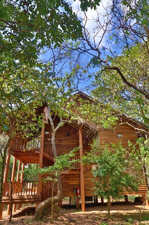 A wooden cabin with a deck and a covered porch, surrounded by trees. - The Other Corner (Habarana)
