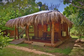 A thatched-roof bungalow with a porch, a small garden, and a view of the surrounding greenery. - The Other Corner (Habarana)