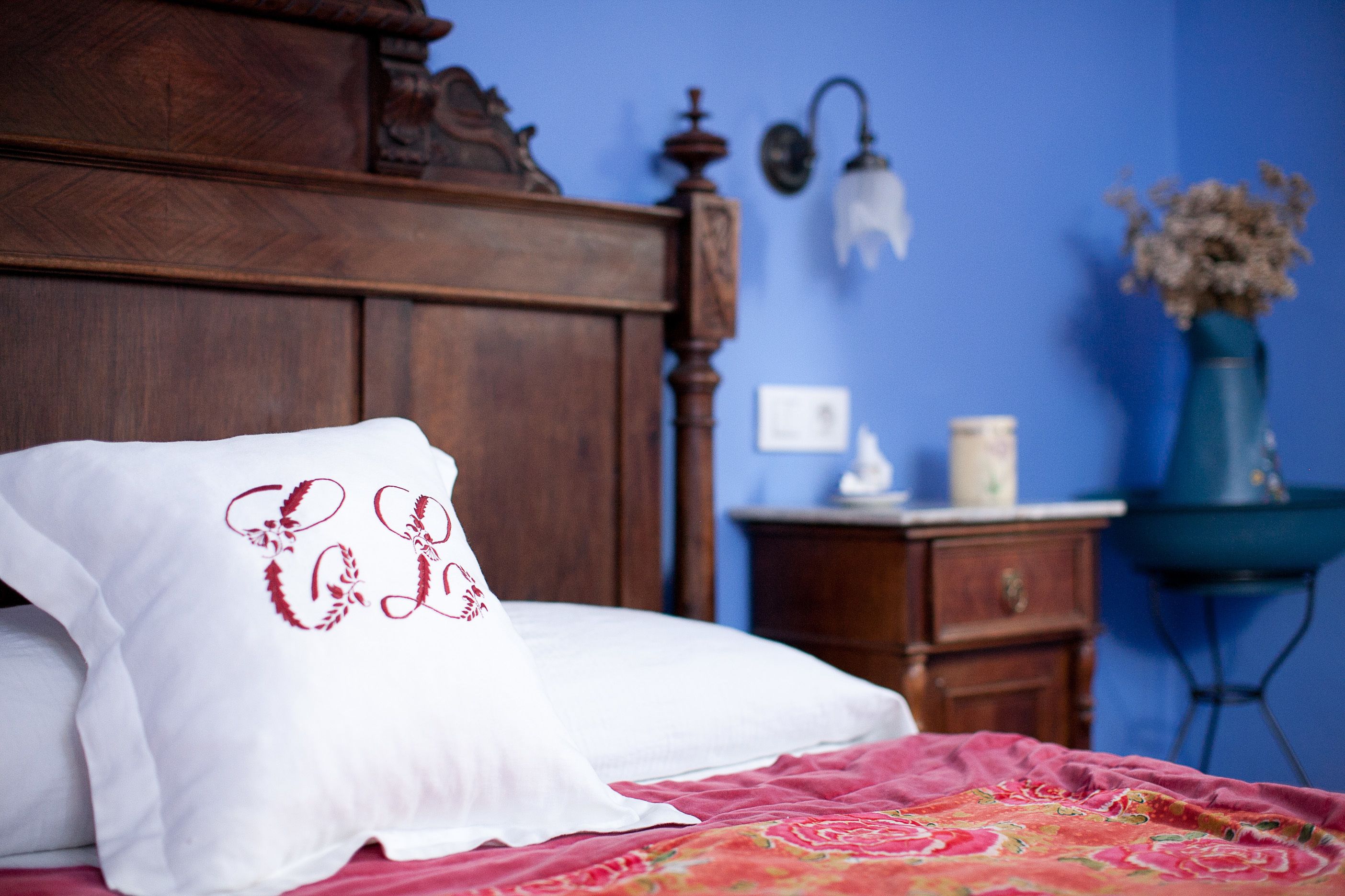 Una cama con almohada blanca y bordado rojo, cabecera de madera, mesita de noche con lámpara y un florero con flores secas.