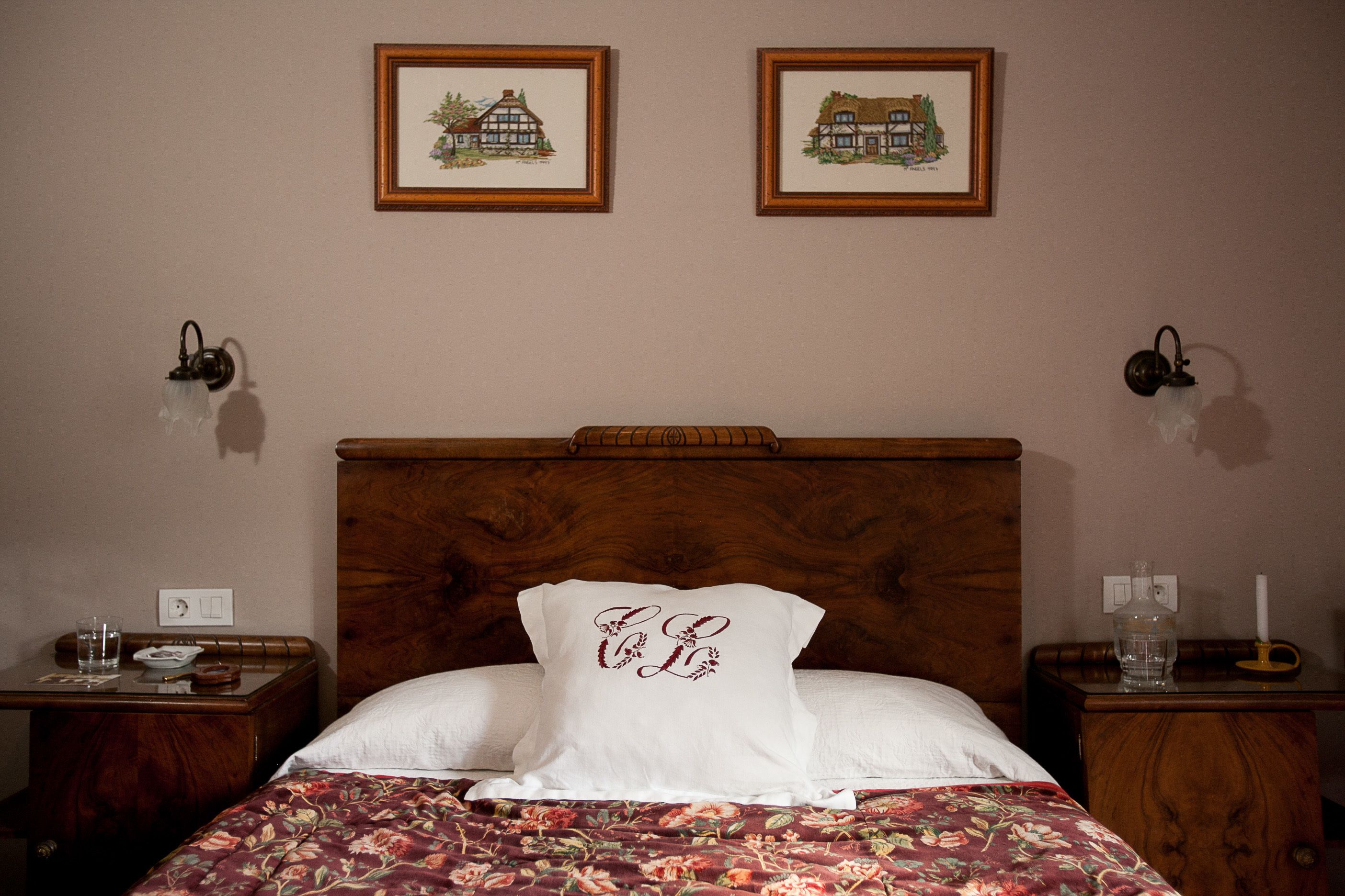 A bedroom with a wooden headboard, floral bedding, two framed pictures on the wall, and bedside tables with lamps and a vase.